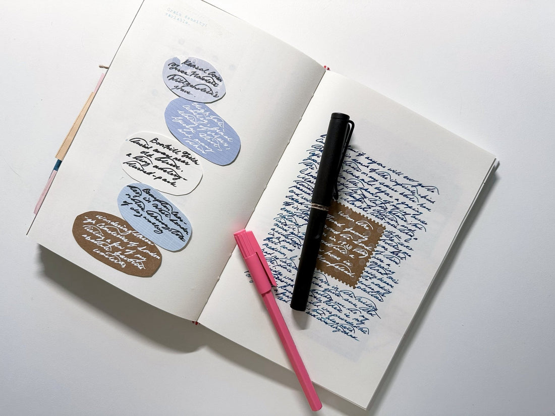 An open Kuura A5 blank notebook, serving as a core diary and life snapshot. The spread is filled with blue ink handwriting, colorful pasted labels, and reflections. A pink brush pen and black pen rest on the pages, ready for journaling.