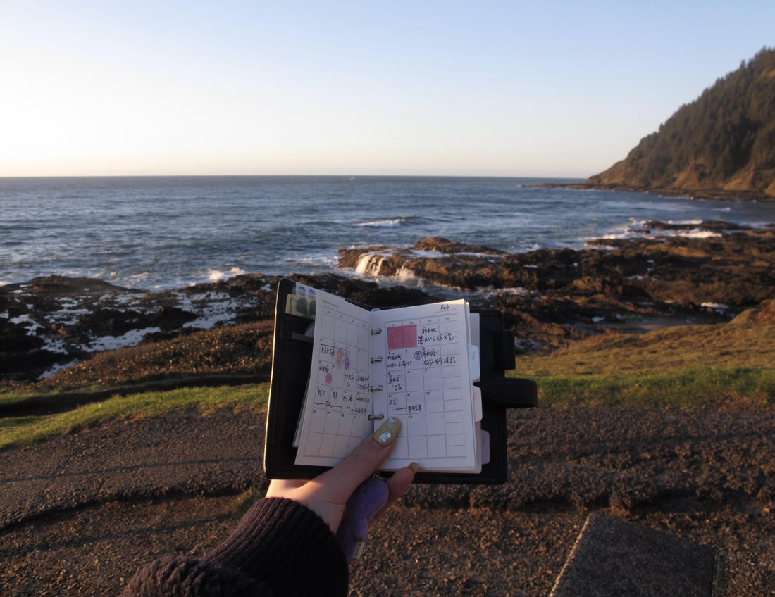 Hand holding an open ring-bound planner with handwritten notes, overlooking a rocky coastline at golden hour