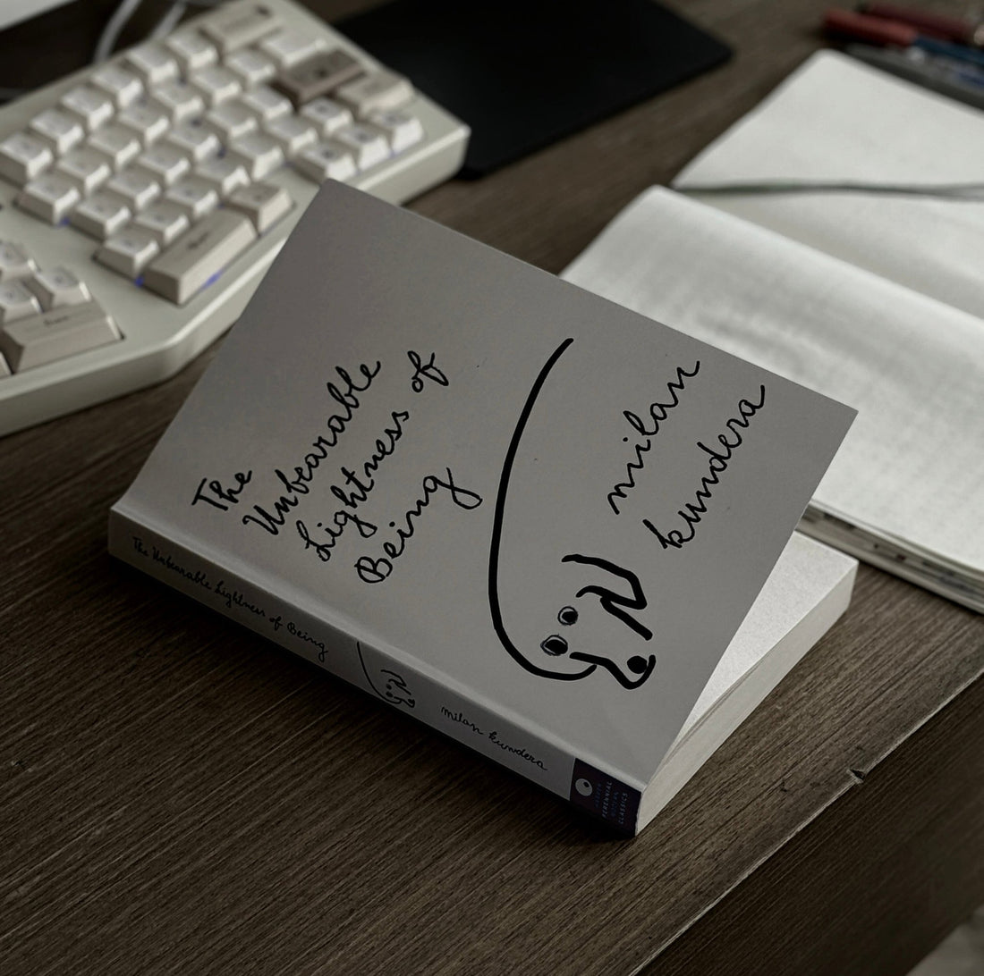 Milan Kundera's The Unbearable Lightness of Being with hand-lettered cover art resting on a wooden desk beside a keyboard and notebook.
