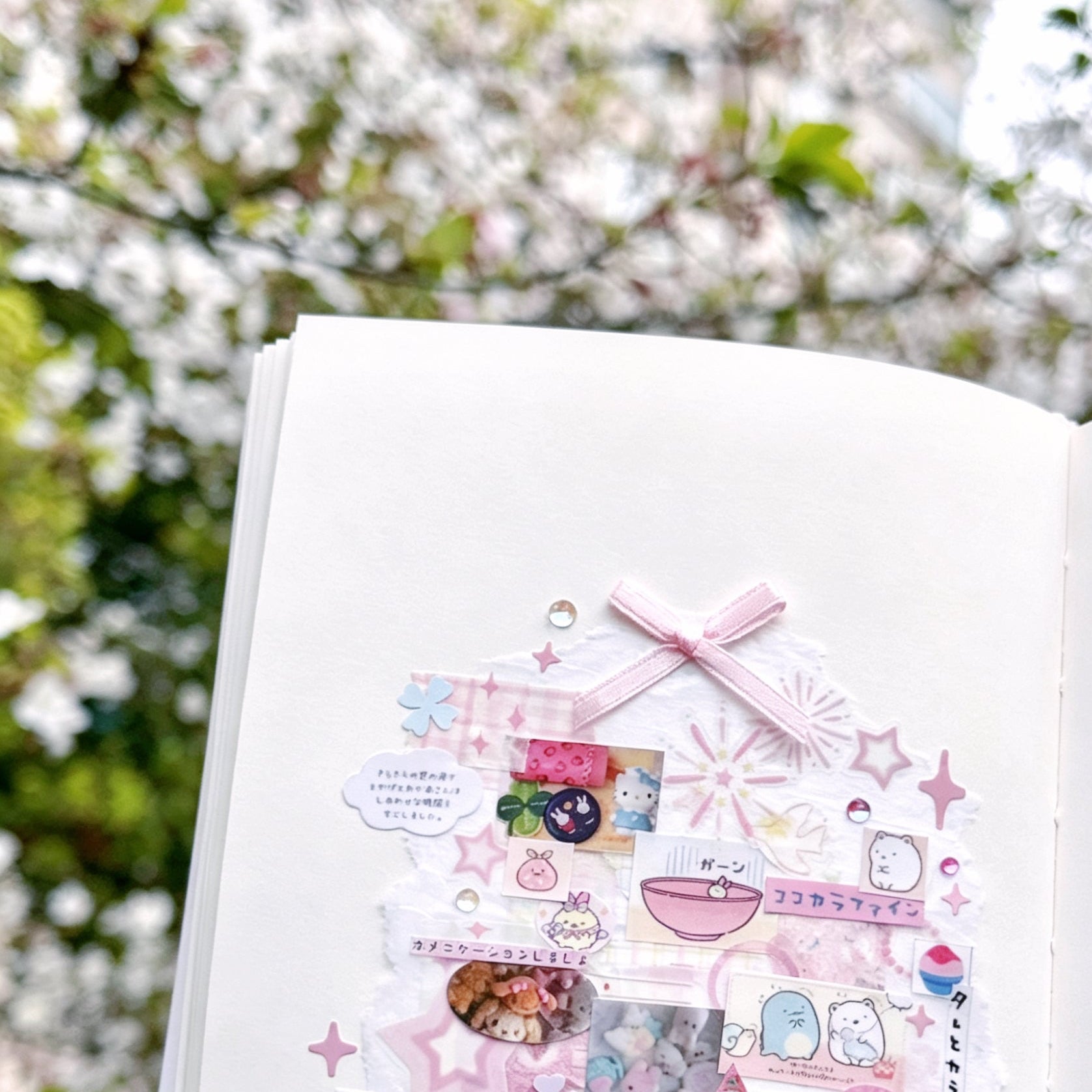 An open Kuura A6 unlined bound notebook is featured against a blurred backdrop of blooming delicate white cherry blossom branches. The page holds an intricate, handmade scrapbooking collage (kawaii aesthetic) composed of layers of torn paper, pink washi tape, adorable character stickers (including Sumikko Gurashi), small printed photos, a tiny pink ribbon bow, Japanese script stickers, and sparkling rhinestone charms, showcasing the creative possibilities of blank pages