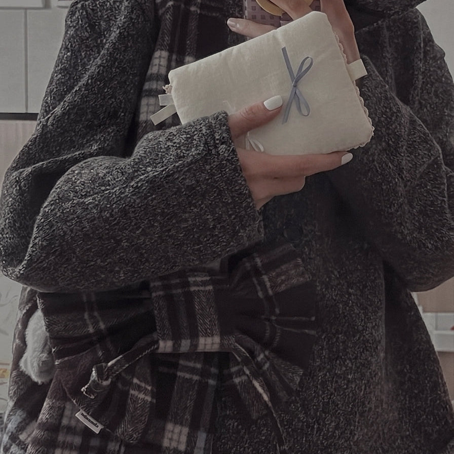 A person wearing a cozy grey herringbone coat with a large plaid bow holding a cream-white kuura fabric journal cover decorated with a light blue ribbon. The image showcases a vintage, academia, or mori-girl style aesthetic, highlighting the journal cover as a stylish fashion accessory.