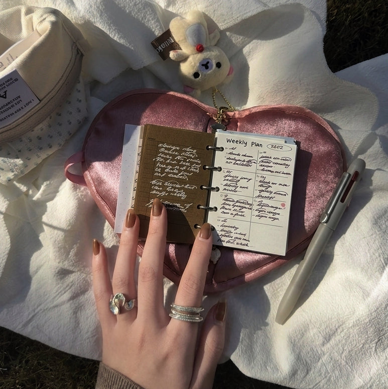 Outdoors in bright sunlight, a hand with brown nail polish and rings is flipping through a kuura 6-ring mini binder journal, showing a 'Weekly Plan' insert. The notebook rests on a pink satin heart-shaped pouch, surrounded by a white knit blanket, a beige pencil case, a multi-color pen, and a cute bear bag charm.