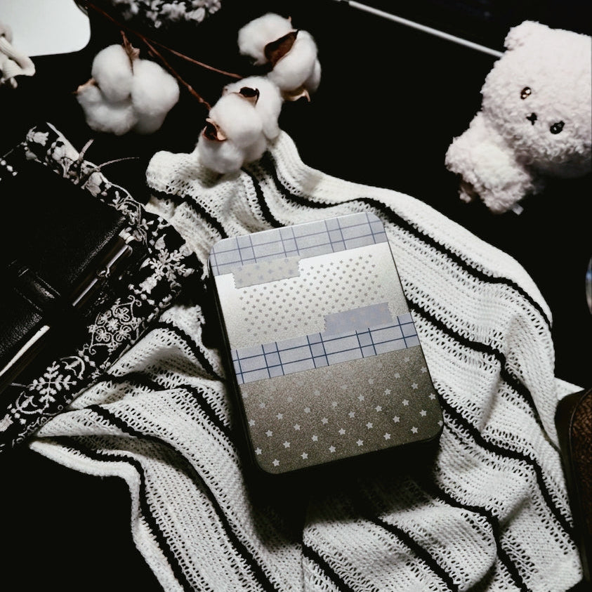 A cozy desk setup featuring a kuura tin storage tin decorated with washi tape patterns (plaid, polka dots, stars) resting on a white and black striped knit blanket. The background includes cotton stems, a black patterned journal, a brass wax seal stamp, a cute bear plushie, and a glowing digital clock, showcasing washi tape collection and desk organization.