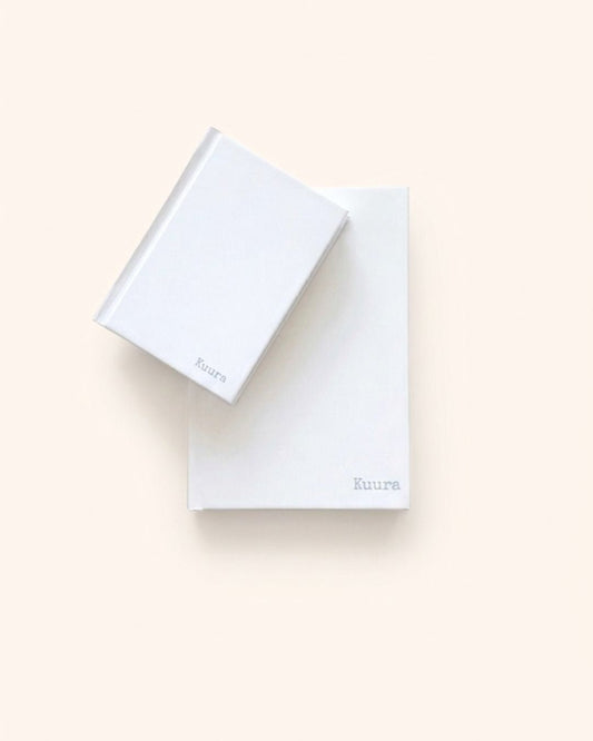 Minimalist flat lay photograph of two closed Kuura Unlined Hardcover Notebooks stacked—one A5 and one A6 size—showcasing the size options. Both durable journals feature the debossed brand logo on the sturdy white casing, perfect for aesthetic stationery planning and gift ideas