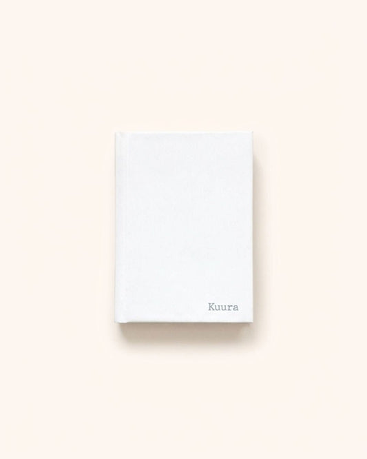 Flat lay photograph of the closed elegant white Kuura A5 & A6 Unlined Hardcover Notebook, showcasing its sturdy casing and delicate debossed brand logo in the bottom right corner. A minimalist and durable journal ideal for an aesthetic sanctuary for thoughts and dreams