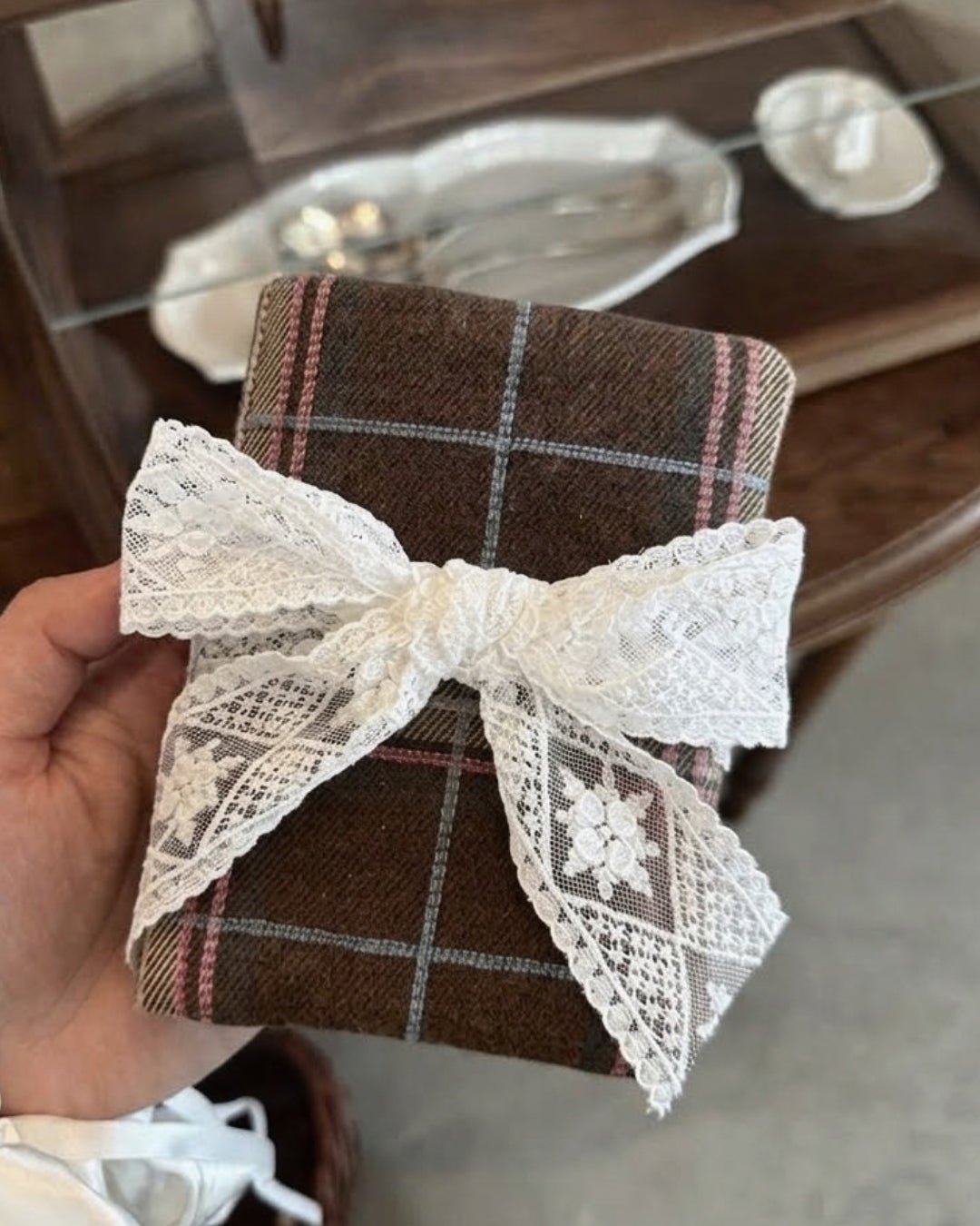 Kuura Museum Date A5 & A6 Journal & Notebook Cover in Brown Plaid, held in hand with its ivory lace bow by an antique display cabinet. Journaling gift idea