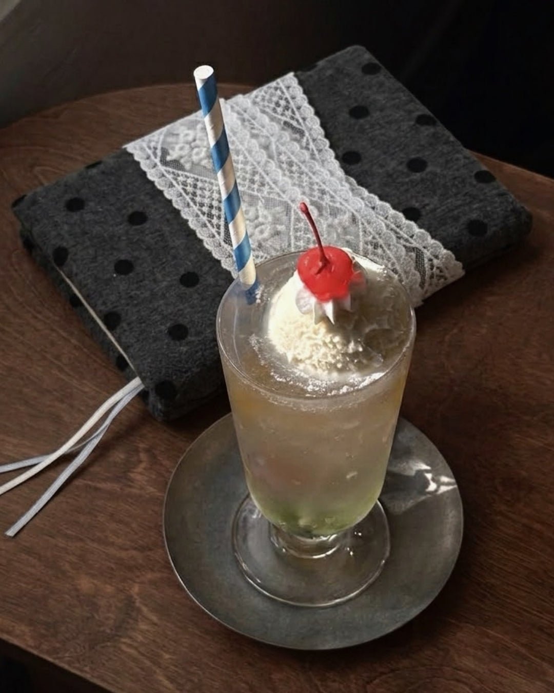Kuura Polka Dot A5 & A6 Journal & Notebook Cover in Charcoal, black dots with white lace trim, on a wooden table beside an ice cream float. Cafe journaling